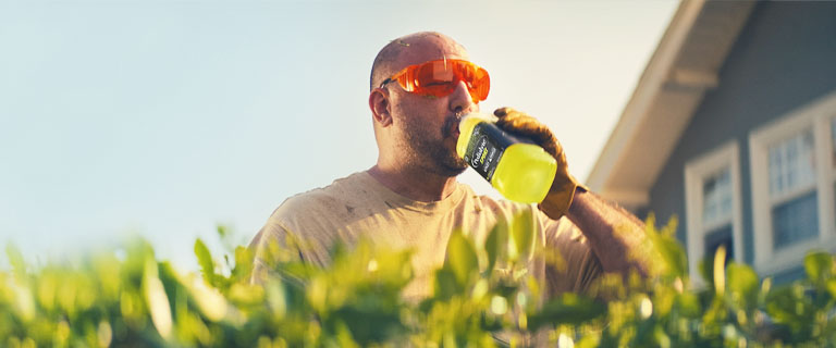 A sweaty man trimming hedges in the heat takes a break to drink from a liter of Pedialyte® Sport for hydration.