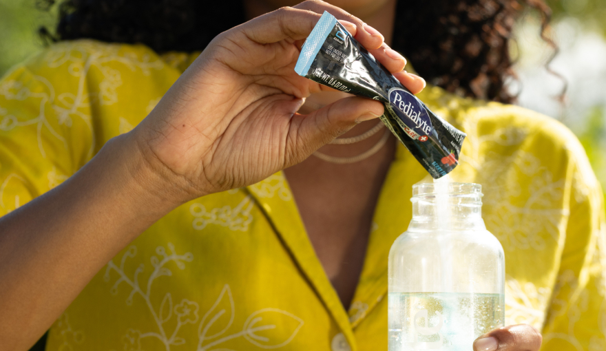 Seasonal homepage image, woman pouring a berry frost flavored Pedialyte advanced care powder pack into water bottle for on-the-go hydration.